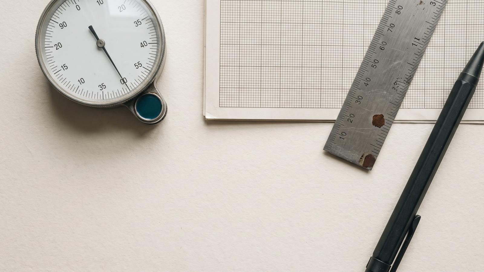 Overhead view of an analogue dial gauge, a sheet of graph paper, a steel rule and a mechanical pencil arranged on a pale paper surface.