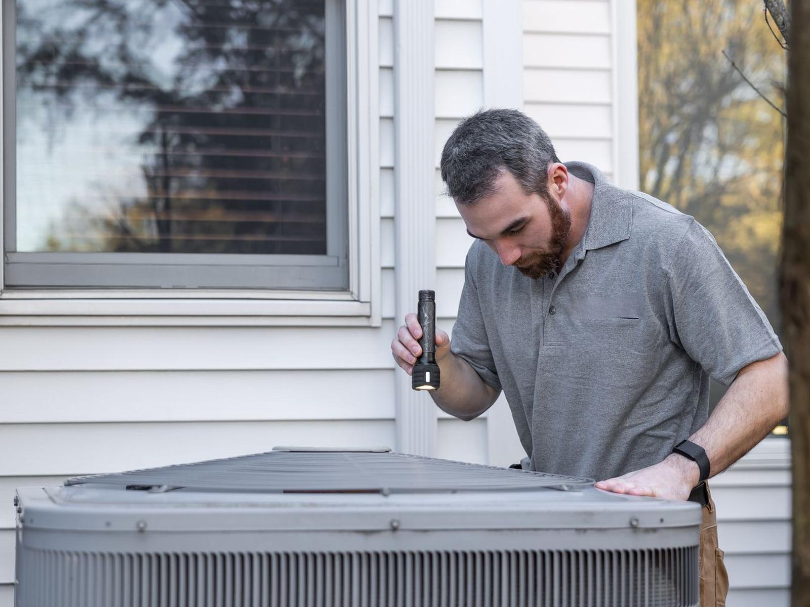 A service technician inspecting a residential outdoor condensing unit with a flashlight.